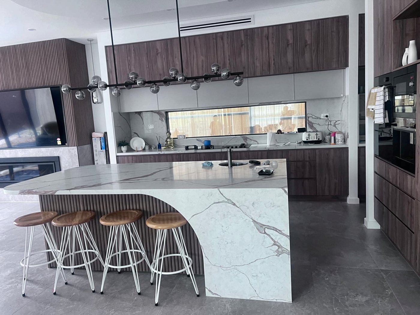A kitchen with a curved marble waterfall island, timber-look cabinetry and a row of bar stools